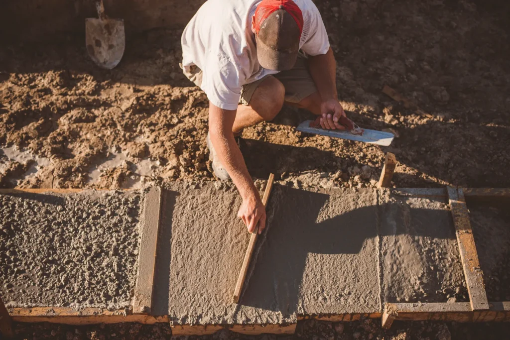 A person kneeling on the ground, smoothing wet concrete in a construction site.