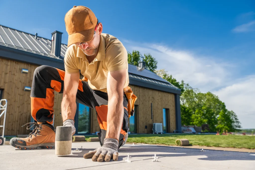 A construction worker in a cap and gloves is kneeling on a construction site, using a tool to secure materials on a sunny day. A modern house is visible in the background.
